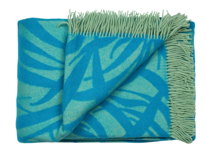 Et foldet turkis og lysegrønt tæppe med en frynset kant og et abstrakt bladmønster.