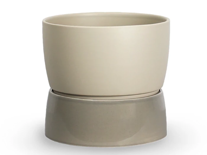 En beige keramikpotte står oven på en lidt bredere, grå keramikunderkop på en hvid baggrund.