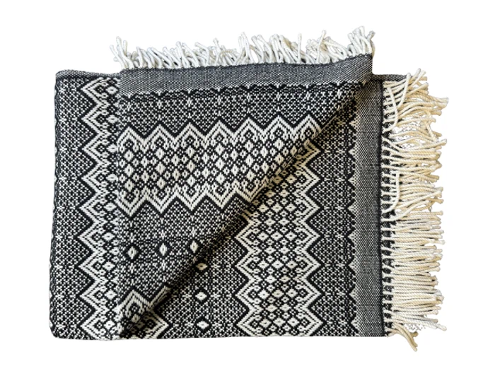 Et foldet sort og hvidt vævet tæppe med geometriske mønstre og frynsede kanter, vist på en hvid baggrund.
