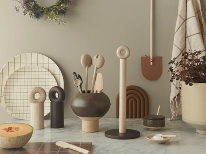 En moderne køkkenscene med træredskaber i en vase, keramiske beholdere, en udskåret melon, tallerkener og minimalistisk indretning på en marmorbordplade mod en beige væg.