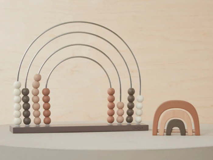 En regnbueformet kugleramme med neutralfarvede perler ved siden af en lille regnbuestabler i træ i lignende jordfarver, begge på en lysegrå overflade.