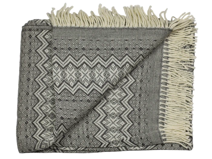 Et foldet gråt og hvidt vævet tæppe med et geometrisk zigzagmønster og frynsede kanter, delvist vendt i det ene hjørne.