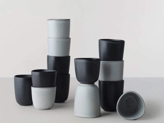 En gruppe minimalistiske keramiske kopper i sort og lysegrå, stablet i forskellige arrangementer på en neutral overflade.