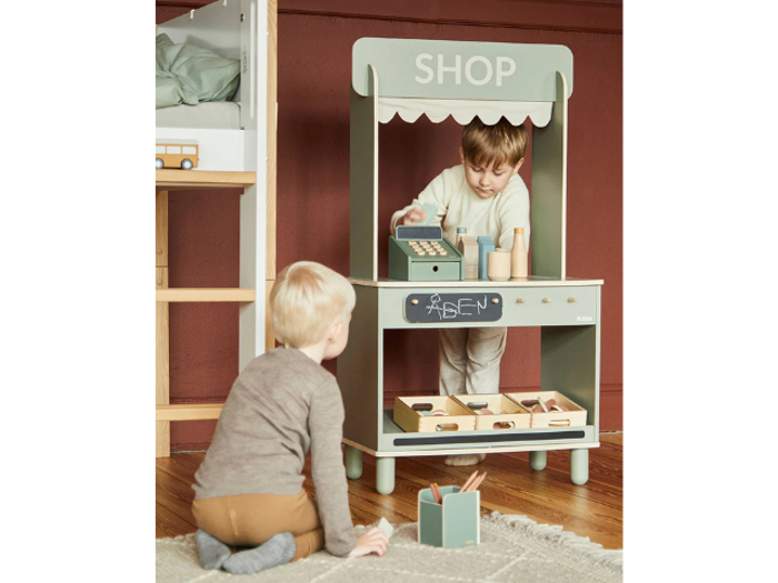 To små børn leger ved en legetøjsbutik i træ. Den ene står bag disken, mens den anden sidder på gulvet med front mod standen. Legetøjsartikler og et kasseapparat er synlige.