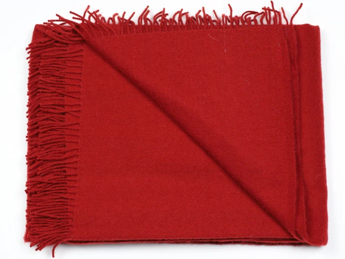 Et foldet rødt uldtørklæde med frynser langs den ene kant, sirligt arrangeret på en hvid baggrund.