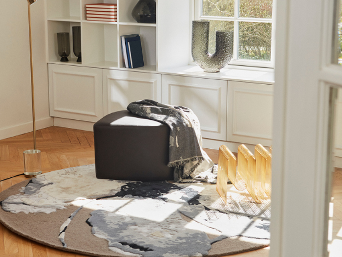 En hyggelig, solbeskinnet læsekrog med en sort skammel, grå plaid, rundt abstrakt tæppe, bogreol og moderne indretning nær et stort vindue.