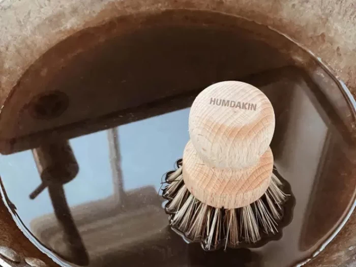 En opvaskebørste af træ mærket Humdakin flyder i en mørk væske inde i en rund beholder eller vask.