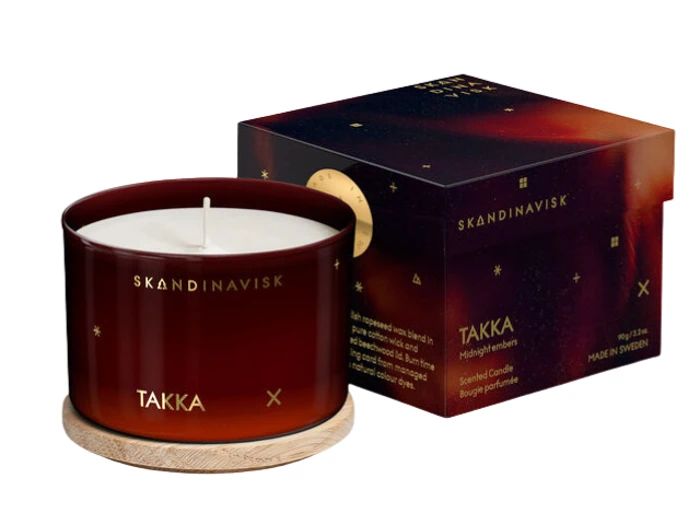 Et rødt SKANDINAVISK TAKKA duftlys med træfod er placeret ved siden af den matchende dekorative æske med gulddetaljer og midnatsinspireret design.