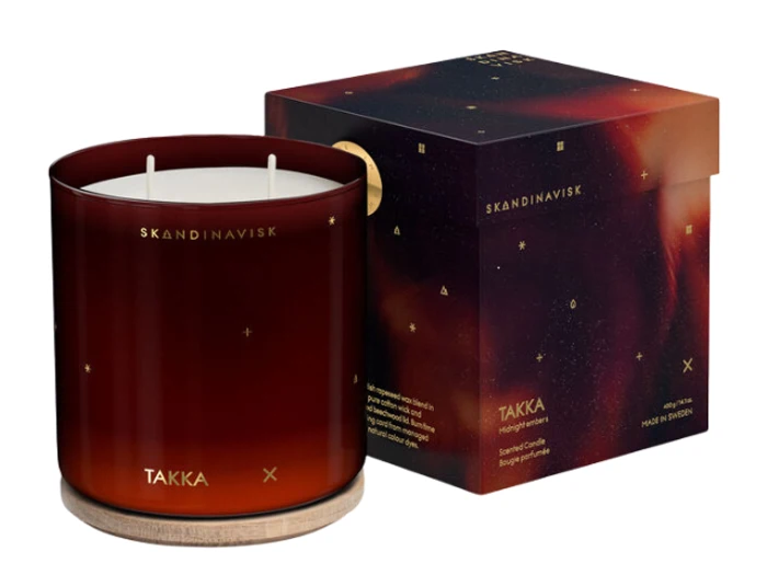 Et mørkt ravfarvet glaslys mærket SKANDINAVISK TAKKA med en træfod, placeret ved siden af sin matchende mønstrede æske.