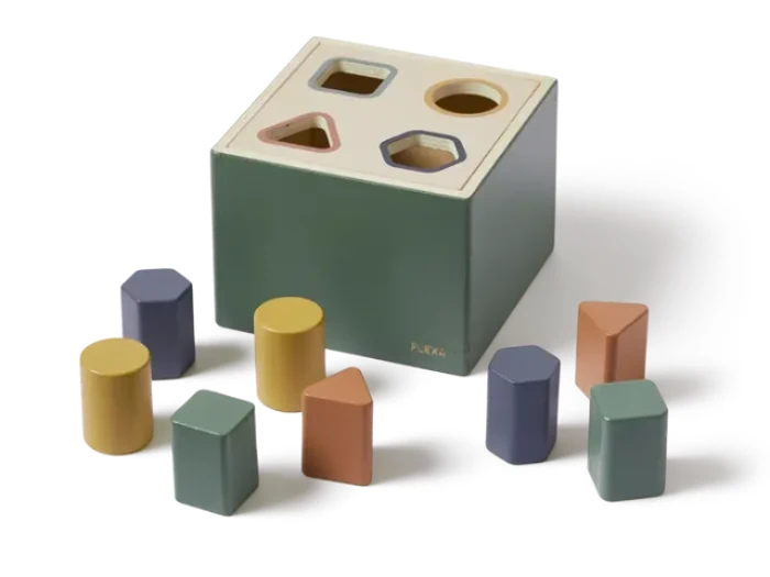 En grøn formsorteringskasse med fire huller på toppen og otte farverige geometriske blokke, herunder cylindre, trekanter og rektangler, spredt rundt om den.