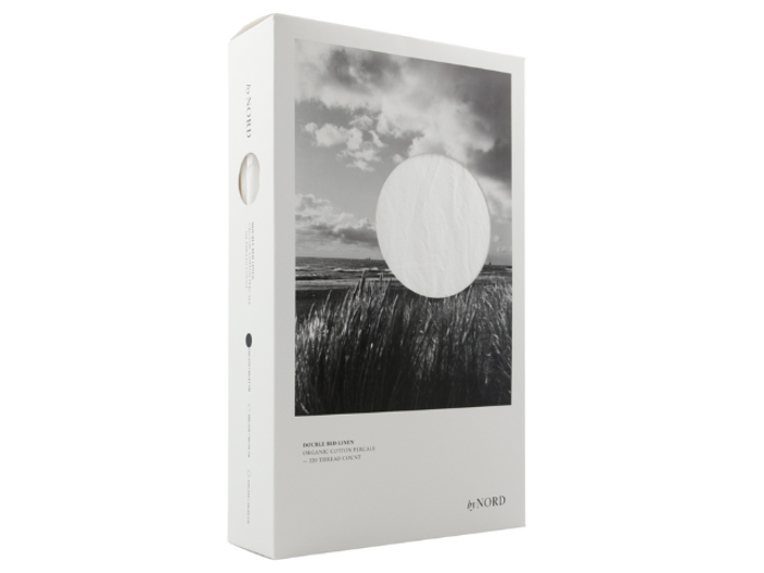 En hvid æske med et sort-hvidt foto af en mark og en himmel, som indeholder et sengesæt af mærket byNORD. Emballagen er minimalistisk og elegant.
