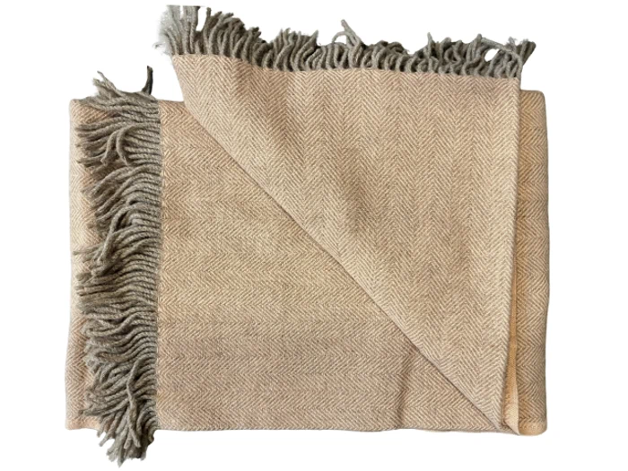 Et foldet beige tæppe med et diskret sildebensmønster og grå frynsekanter, vist delvist udfoldet i det ene hjørne.