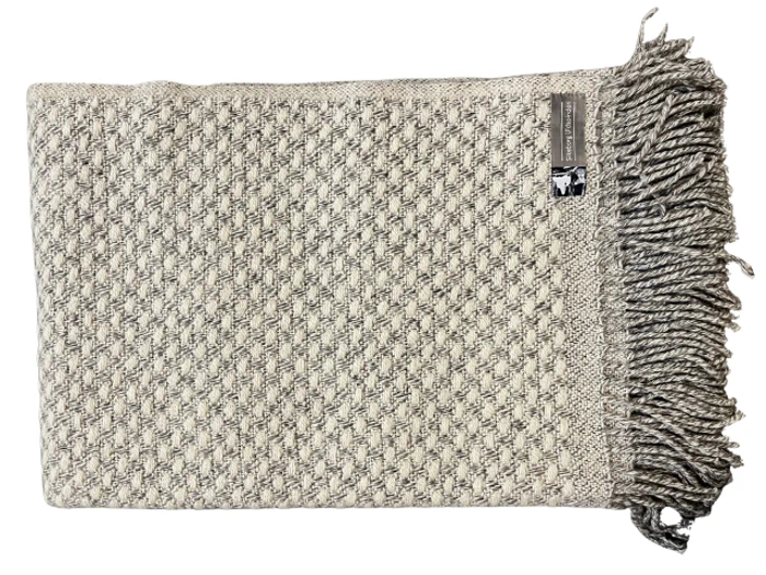 Et foldet, beige og gråt vævet tæppe med et struktureret diamantmønster og en frynset kant på den ene side. En lille etiket er fastgjort nær kanten.