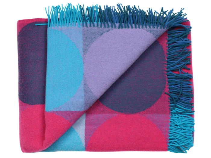 Et foldet uldtæppe med et stort geometrisk mønster i pink, lilla og blå og blågrønne frynser langs den ene kant.