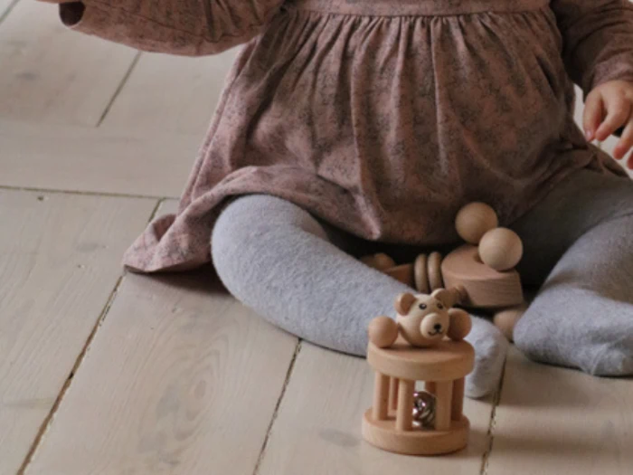 Et lille barn iført grå strømpebukser og en lyserød kjole sidder på et trægulv og leger med trælegetøj, bl.a. en lille bjørn i et bur.