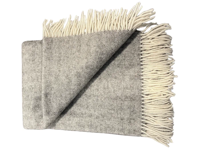 Et foldet gråt sildebensmønstret tæppe med cremefarvede frynser på kanterne, lagt på en hvid baggrund.