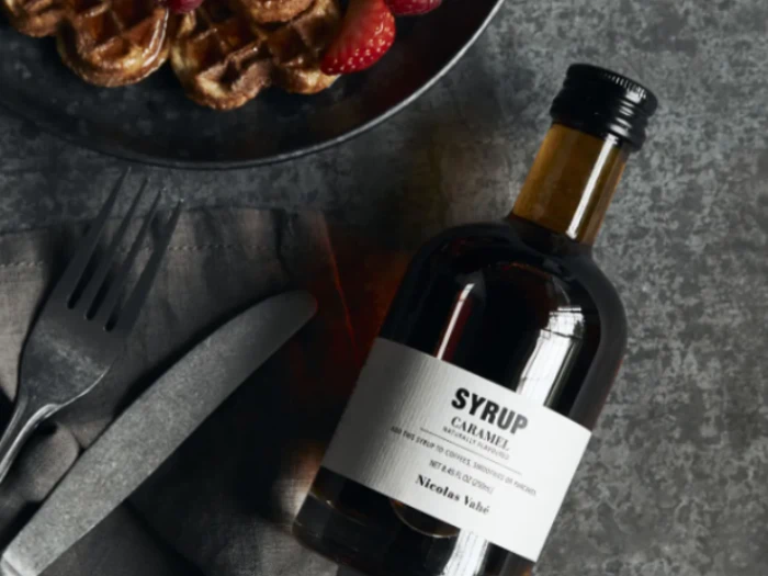 En flaske karamelsirup ligger ved siden af en gaffel og en kniv på en serviet, og en tallerken med vafler og jordbær er delvist synlig ovenover.