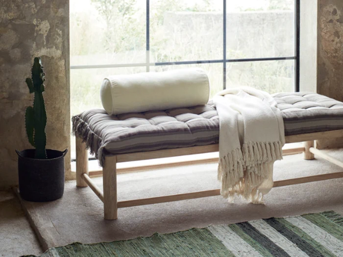 En polstret træbænk med en sammenrullet pude og et frynset tæppe står ved et stort vindue; en kaktus i potte står ved siden af på gulvet, og et grønt tæppe er delvist synligt.