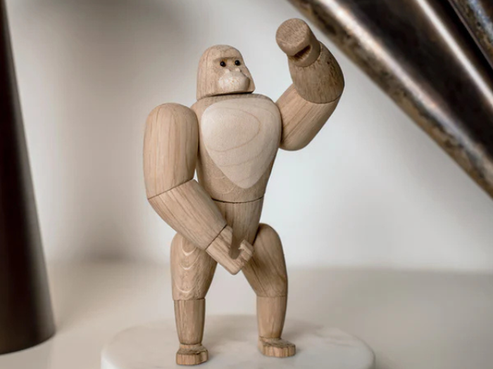 En gorillafigur i træ står på en rund, hvid base med en løftet arm og et neutralt ansigtsudtryk.