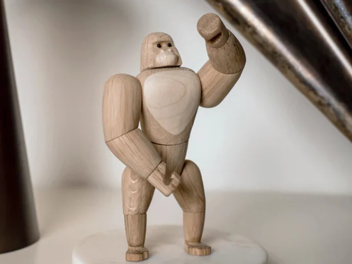 En gorillafigur i træ står på en rund, hvid base med en løftet arm og et neutralt ansigtsudtryk.