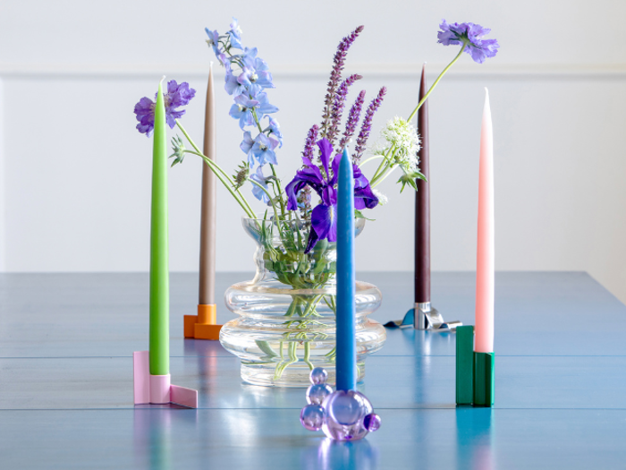 En glasvase med lilla og blå blomster står på et blåt bord omgivet af farverige stearinlys i moderne geometriske stager.