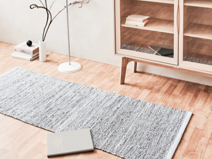 Et gråt vævet tæppe på et lyst trægulv ved siden af et skab med bøger i og en gulvlampe med en tynd gren som dekoration i nærheden. En åben bog ligger på gulvet.
