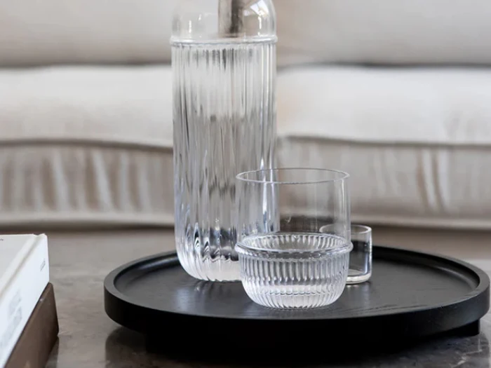 En vandkaraffel i riflet glas og to matchende glas står på en sort, rund bakke, der er placeret på et bord med en lys sofa i baggrunden.