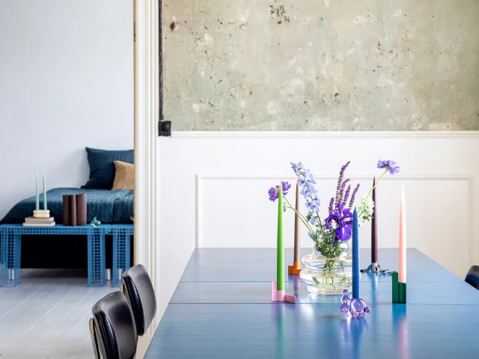 Et moderne spisebord med farverige stearinlys og lilla blomster i en glasvase; et minimalistisk soveværelse med en blå seng ses i baggrunden.