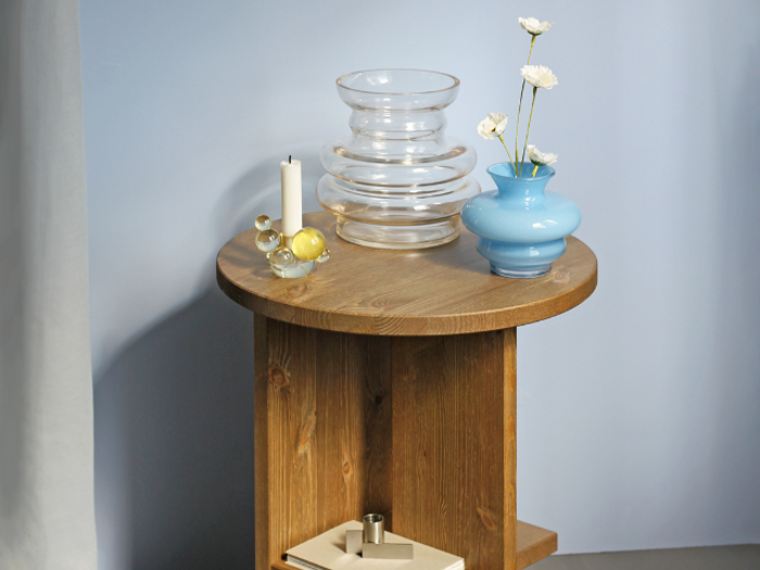 På et rundt sidebord af træ står en blå vase med hvide blomster, en bølget klar vase, et hvidt stearinlys og glaspynt; på den nederste hylde står bøger og en lille ting.