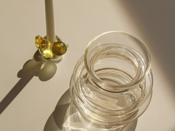 En klar glasvase kaster refleksioner på en lys overflade ved siden af en glasrører med runde gule detaljer, begge oplyst af sollys.