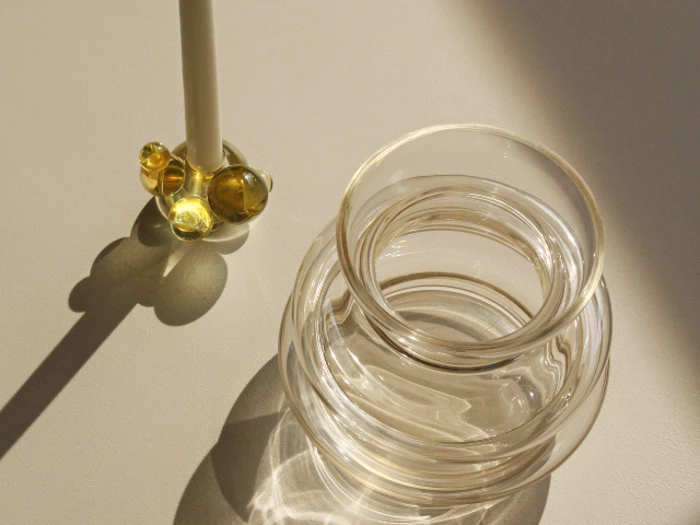 En klar glasvase kaster refleksioner på en lys overflade ved siden af en glasrører med runde gule detaljer, begge oplyst af sollys.