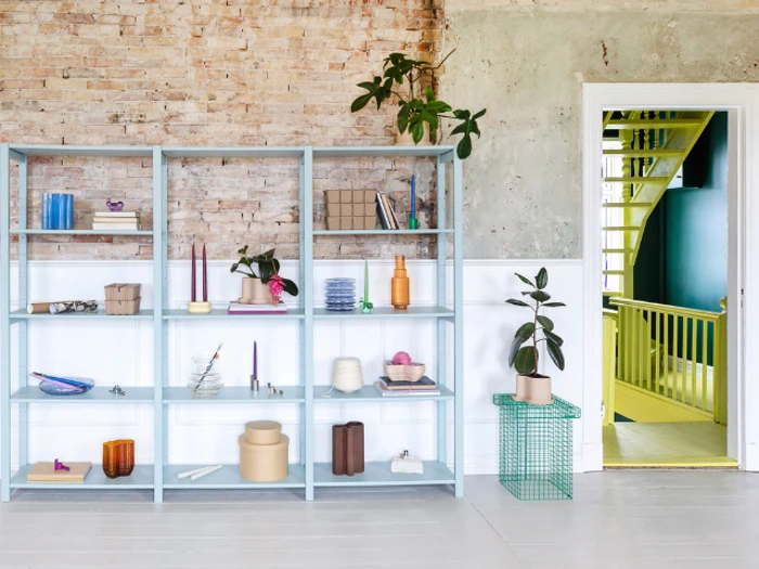 En lyseblå reol med pyntegenstande står op ad en rustik murstens- og gipsvæg ved siden af en døråbning, der afslører en lysegul trappe.