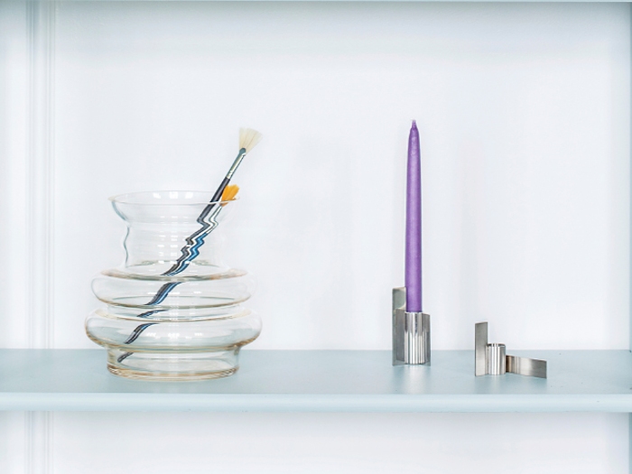 En klar, bølget glasvase med pensler indeni står ved siden af et højt lilla lys i en moderne sølvholder på en lyseblå hylde.