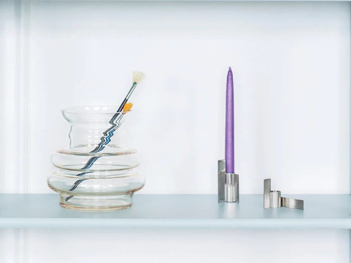 En klar, bølget glasvase med pensler indeni står ved siden af et højt lilla lys i en moderne sølvholder på en lyseblå hylde.