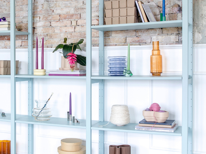 Lyseblå hylder med bøger, stearinlys, planter, garn, glas og pyntegenstande mod en hvid panel- og murstensvæg.