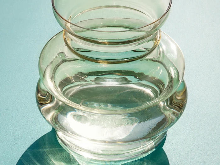 En klar glasvase med afrundede kanter står på en turkis overflade og kaster skygger og refleksioner i sollyset.