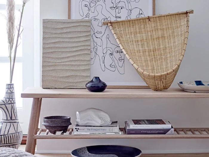 Et konsolbord i træ med kunst, en rottingkurv, vaser, bøger og keramikudsmykning i en minimalistisk, moderne bolig.