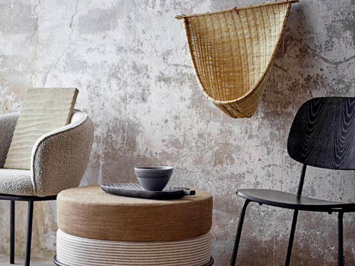 Et hyggeligt opholdsområde med en tekstureret lænestol, en sort stol, en rund beige ottoman med skåle på toppen og en flettet kurv, der hænger på en forvitret betonvæg.