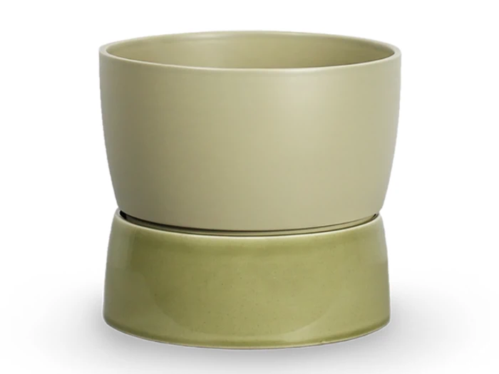 En beige keramikpotte står oven på en omvendt, lidt mørkere grøn underkop, begge med en glat, mat finish på en hvid baggrund.