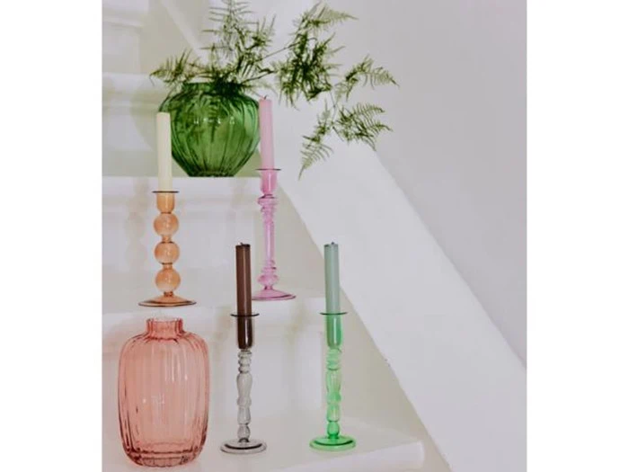 Farverige glaslysestager med stearinlys og to vaser, en grøn med bregneblade og en lyserød, arrangeret på en hvid trappe.