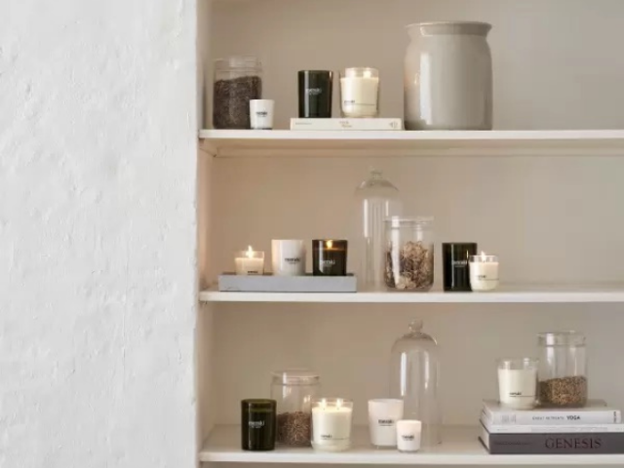 Tre hvide hylder med tændte og utændte stearinlys i glaskrukker, bøger, en stor keramikvase og krukker fyldt med tørret indhold mod en hvid væg.