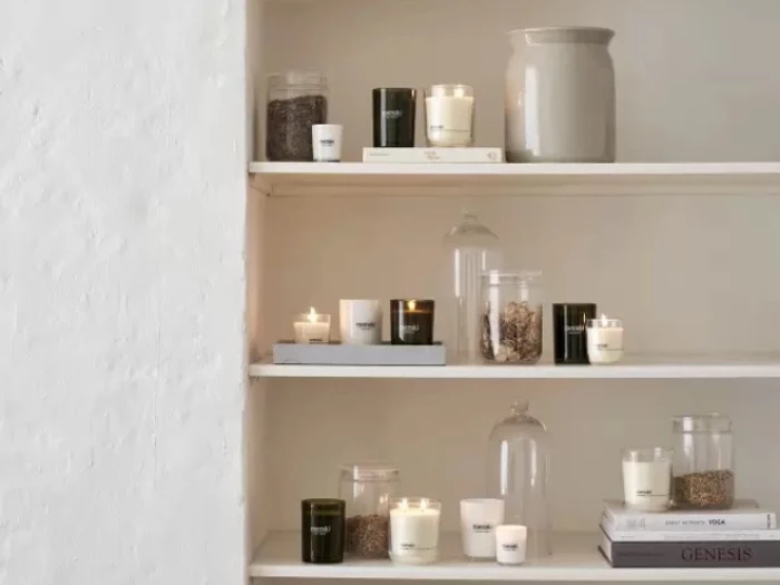 Tre hvide hylder med tændte og utændte stearinlys i glaskrukker, bøger, en stor keramikvase og krukker fyldt med tørret indhold mod en hvid væg.