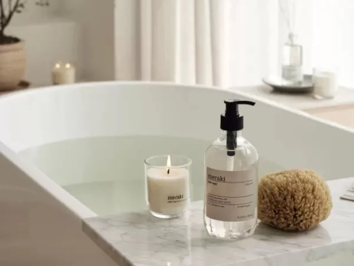 Et badekar fyldt med vand står ved siden af en marmorkant med et tændt stearinlys, en klar sæbedispenser og en naturlig svamp. Baggrunden har blød belysning og minimal indretning.