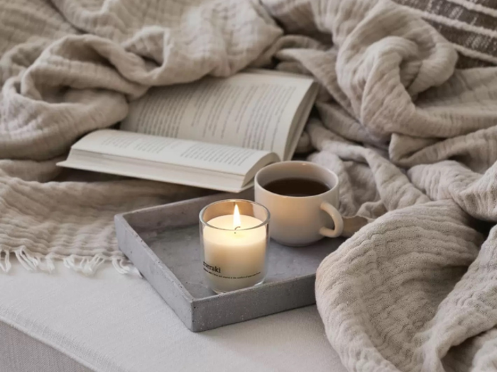 Et tændt stearinlys og en kop kaffe står på en bakke ved siden af en åben bog på en hyggelig, beigefarvet seng med tæppe.