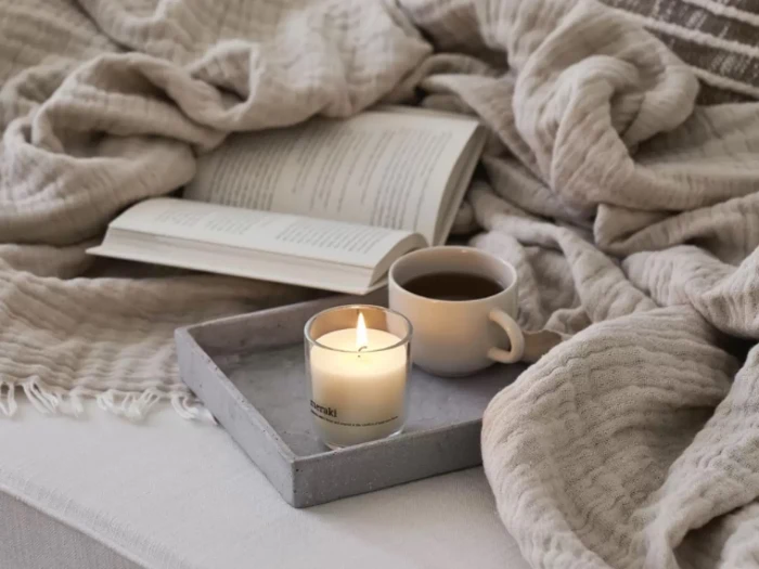 Et tændt stearinlys og en kop kaffe står på en bakke ved siden af en åben bog på en hyggelig, beigefarvet seng med tæppe.