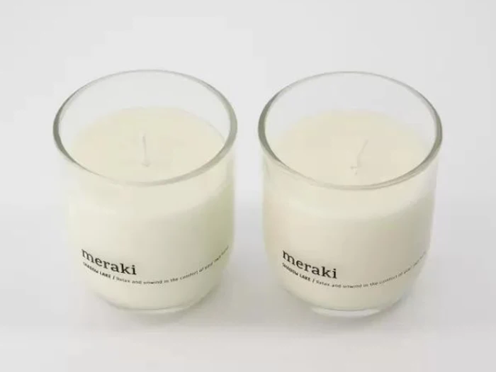 To hvide lys i klare glasbeholdere, hver mærket meraki, placeret side om side på en almindelig hvid overflade.