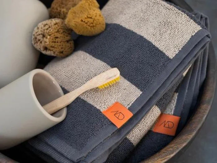 Et stribet håndklæde med et orange mærke, en bambustandbørste, natursvampe og en hvid kop er arrangeret sammen i en skål.