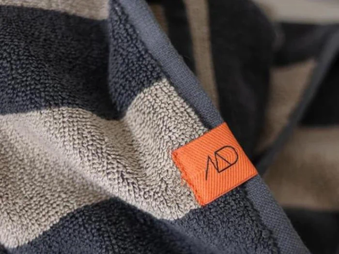 Nærbillede af et stribet håndklæde med marineblå og lysegrå bånd og et orange mærke med et minimalistisk sort AD-logo.