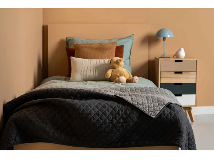 En pænt redt enkeltseng med lagdelte puder og en bamse ved siden af et lille natbord med skuffer, en blå lampe og en dekorativ fugl i et rum med varme farver.