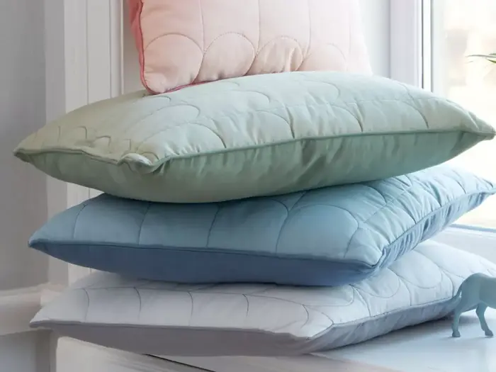 Tre pænt stablede puder i pastelfarver - grøn, blå og hvid - med en lyserød pude delvist synlig bag dem i nærheden af et vindue.